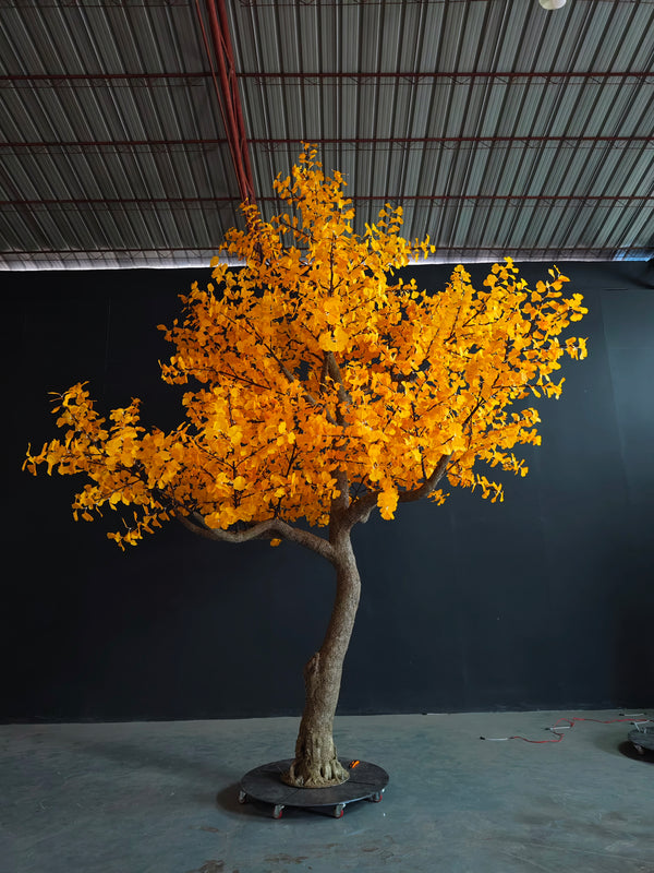 An artificial ginkgo tree with bright yellow leaves and a textured brown trunk, displayed on a black wheeled base in an indoor warehouse setting.