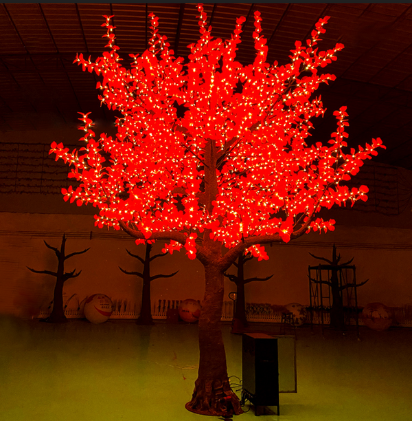 A decorative artificial tree with glowing red lights throughout the branches, photographed indoors against a dark ceiling and floor with other dark tree silhouettes in the background.