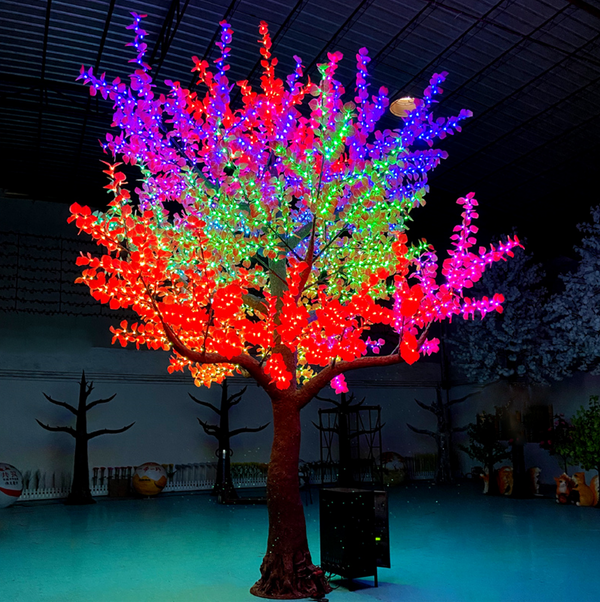 A large decorative tree with glowing multicolor LED lights in red, orange, yellow, green, blue, and purple, set in a dark indoor event space with other illuminated trees in the background.