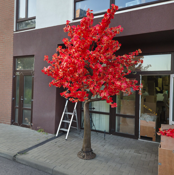 A decorative artificial cherry blossom tree with bright red flowers and a brown trunk, displayed outdoors on a paved sidewalk in front of a building with glass doors and a ladder nearby.