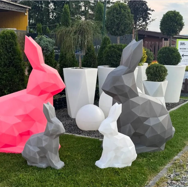 Geometric rabbit sculptures in various colors on grass with plants and a building in the background
