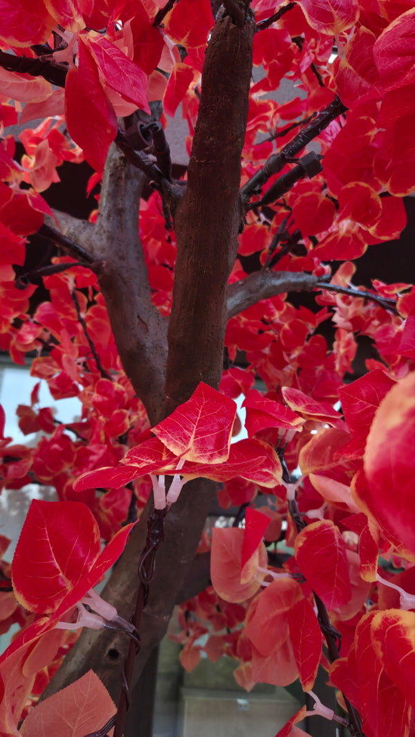 A close-up of a red artificial tree with bright red leaves and dark brown branches, photographed tightly with some leaves blurred in the foreground.