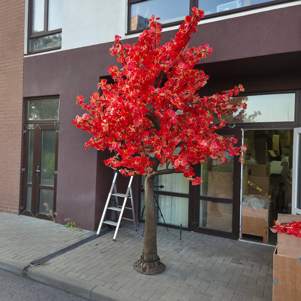 An artificial flowering tree with bright red blossoms and a brown trunk, displayed outdoors on a paved sidewalk in front of a building with glass doors and windows.