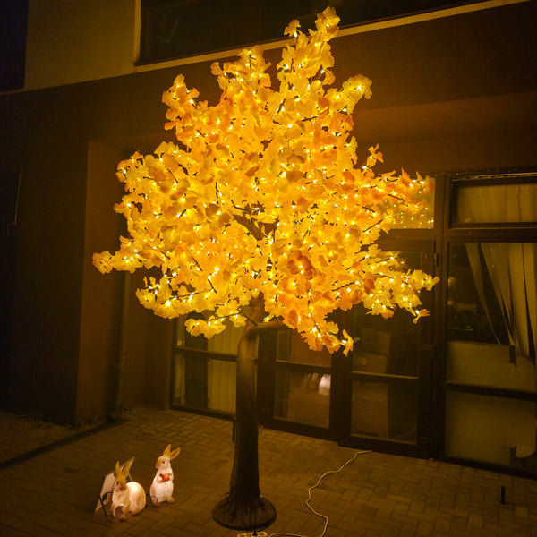 A large artificial flowering tree with glowing warm yellow-orange lights, displayed outdoors at night in front of a building with dark-framed windows. Two small rabbit figurines sit on the ground nearby.
