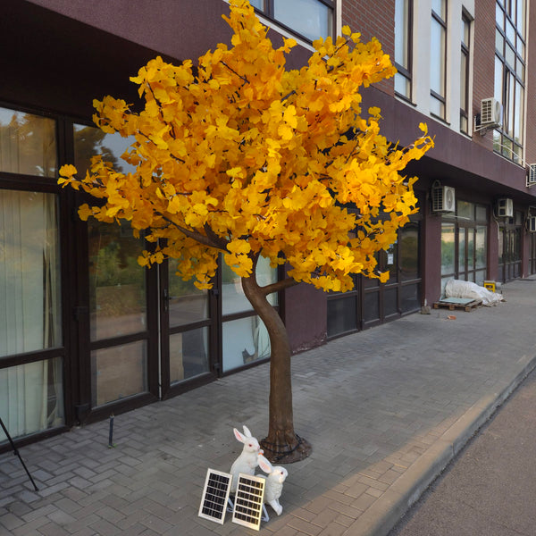 An artificial ginkgo tree with bright yellow leaves and a brown trunk, planted on a paved sidewalk in front of a modern building. Two small white rabbit decorations and two small solar panels are placed at the base of the tree.