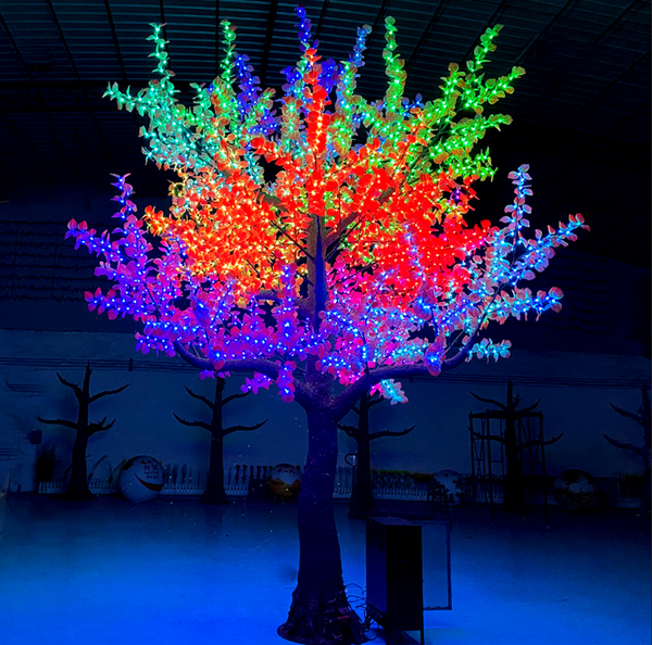 A large artificial LED light tree with a dark brown trunk and branches illuminated in a vibrant rainbow of colors including red, orange, yellow, green, blue, and purple, set against a dark indoor background.
