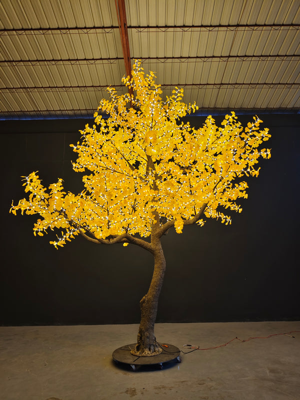 An artificial decorative tree with bright yellow glowing leaves and a brown trunk, mounted on a round black base in an indoor setting with a dark wall and metal ceiling.
