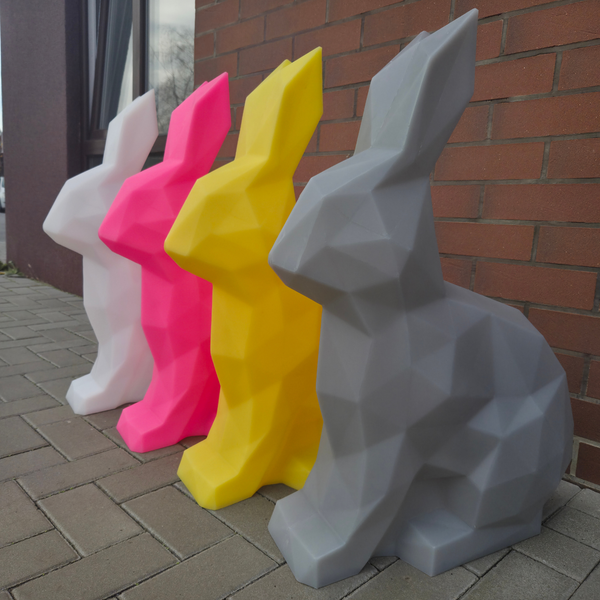 Four geometric rabbit sculptures in different colors (gray, pink, yellow, white) against a brick wall.