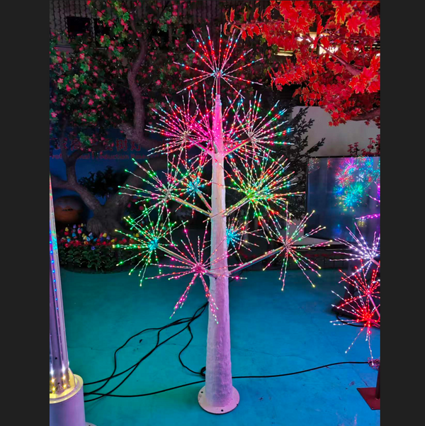 A decorative tree light with a white trunk and branches illuminated by multicolored LED starburst lights, photographed in an outdoor display setting at night.