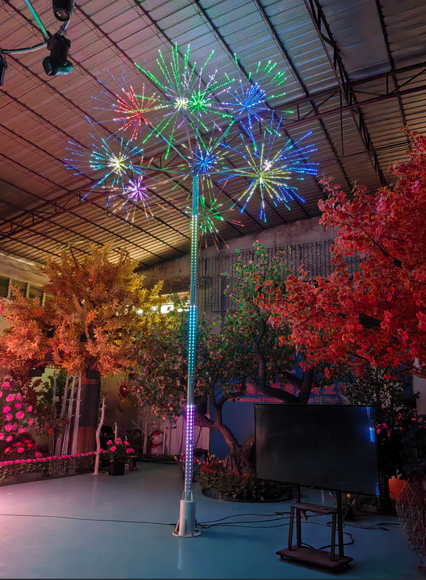 A tall LED decorative tree with a silver pole and multiple colorful starburst lights in blue, green, red, purple, and orange, displayed indoors in a large showroom with artificial trees and a TV stand nearby.