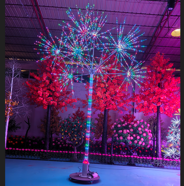 A freestanding LED firework tree with a multicolored lighted trunk and burst-shaped lights, displayed indoors among other decorative trees and floral arrangements under a metal exhibition ceiling.