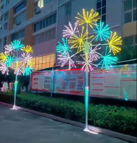 Outdoor LED firework-shaped light trees decorated with bright cyan, yellow, white, and pink lights, set along a sidewalk in front of a building at night.