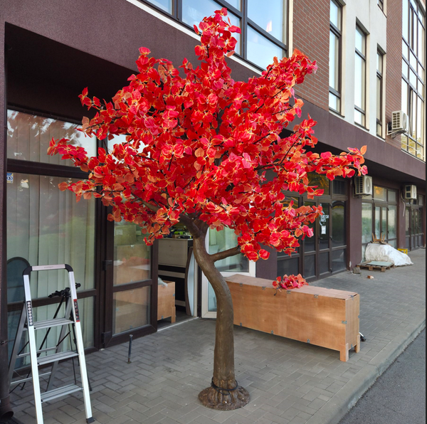 An artificial autumn tree with bright red and orange leaves on a brown trunk, staged on a sidewalk in front of a building with storefront windows.