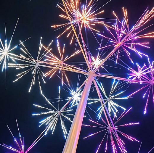 A large outdoor LED light sculpture resembling a dandelion or starburst pattern, illuminated with vibrant purple, pink, orange, and blue lights against a dark night sky.
