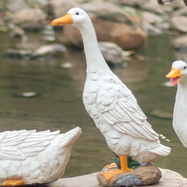 White goose statues with orange beaks on a rock by a body of water.