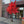 An artificial flowering tree with bright red blossoms and a brown trunk, displayed outdoors on a paved sidewalk in front of a building with glass doors and windows.