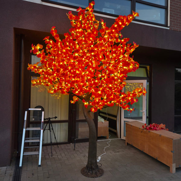 An artificial decorative tree with bright orange and red leaf-shaped lights glowing on a paved outdoor area in front of a building. A white step ladder stands to the left, and the tree is plugged in from a cord on the ground.